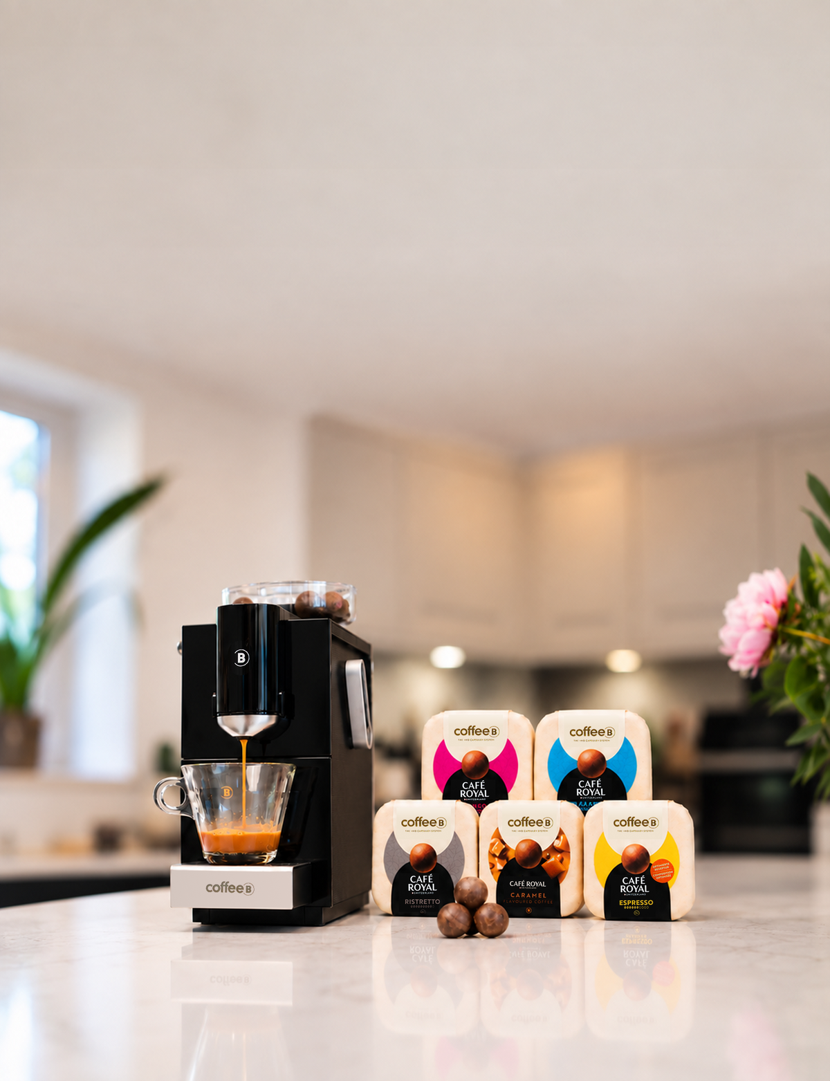 A black CoffeeB Cosmos machine dispensing espresso into a glass coffee cup on a kitchen countertop, with three CoffeeB Coffee Balls displayed, and 5 packs of Coffee Balls lined up beside it showing the Starter Bundle Blends. The background shows a softly blurred modern kitchen interior.